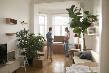 Young married couple carrying boxes into rented flat, cozy room with houseplants, moving into new home after renovation, buying apartment, enjoying relocation. Full length shot - obrazy, fototapety, plakaty