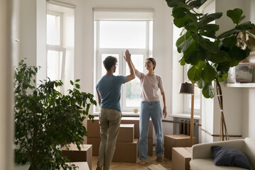 Positive young married couple clapping hands, giving high five over heap of paper relocation boxes...