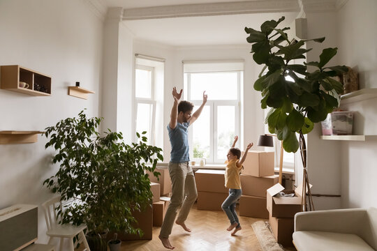 Excited energetic dad and daughter girl enjoying active playtime at home, dancing to music, singing, having fun in new apartment with cardboard boxes stacked in background