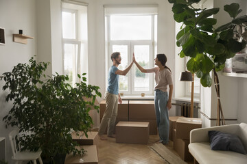 Cheerful satisfied young couple giving high five, clapping deal hands over stack of cardboard boxes in new apartment, moving into cozy flat, celebrating property buying, purchase, unpacking - Powered by Adobe