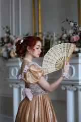 Young red haired woman in ball historic dress and fan in hand in vintage room full of flowers. Portrait of aristicratic woman in historically decorated room. - obrazy, fototapety, plakaty