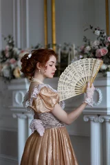 Fotobehang Young red haired woman in ball historic dress and fan in hand in vintage room full of flowers. Portrait of aristicratic woman in historically decorated room. © Альбина Хусаинова