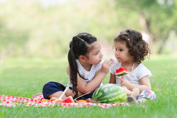 Little Caucasian and Asian girls eating watermelon while picnic at park. Older sister use napkin...