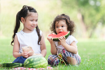 Little Caucasian cute girls eating watermelon while picnic at park. Older sister smiling looking at her adorable sister biting a sliced watermelon. Children sitting on green grass together in summer