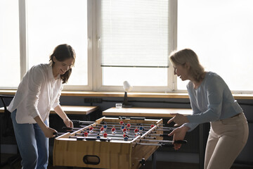 Fototapeta Two happy positive business women of different ages enjoying active break, entertainment in office. Diverse corporate friends playing table football, competing in board soccer game, laughing obraz