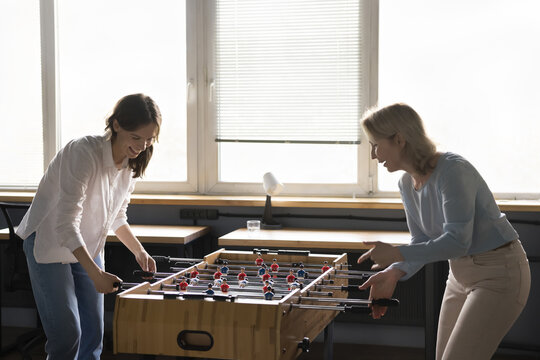 Two happy positive business women of different ages enjoying active break, entertainment in office. Diverse corporate friends playing table football, competing in board soccer game, laughing