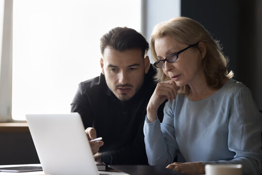 Focused Different Aged Employees, Business Man And Woman Talking At Laptop. Mature Expert In Glasses Showing Online Presentation To Professional Colleague, Speaking At Computer