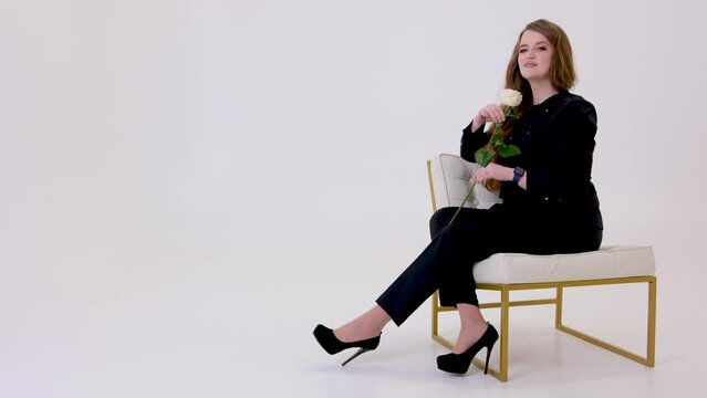 a girl in black sits on a white chair in studio a place for text in her hands a white rose looks forward then a long heel of black platform shoes raises her head at the flower