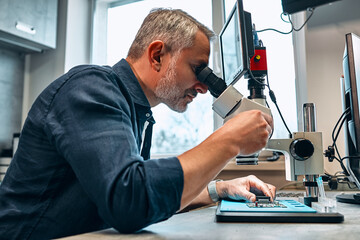 Naklejka na ściany i meble A male engineer looks at a microcircuit through a microscope.Development of microchips, technology.