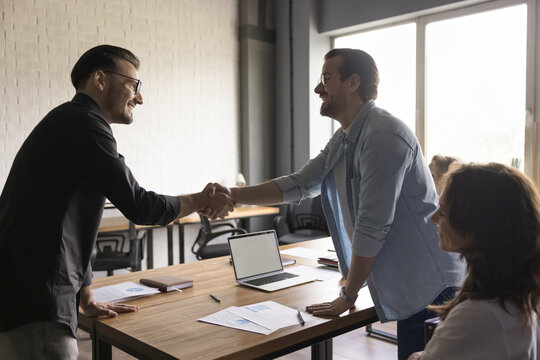 Two positive confident businessmen shaking hands over meeting table after negotiation, closing deal, contract, agreement, smiling. Boss, executive hiring new worker, giving handshake