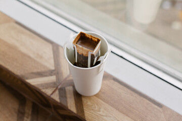 Drip bag in a paper cup on windowsill. Instant freshly brewed alternative drink in mug. Easy brew black coffee method for home, travel or office. cozy trendy lifestyle