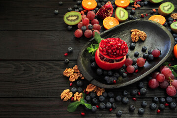 Sliced ​​red, ripe pomegranates are beautifully arranged in a black wooden plate on a black wooden table. Garnished with green mint leaves, fruits, berries, nuts and honey.