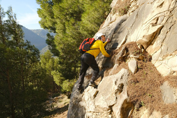tourist in yellow jacket with orange backpack climbs rock, mature man in safety helmet, rock climber, extreme people lifestyle, mountaineering, life insurance, difficulties in hike, extreme sports