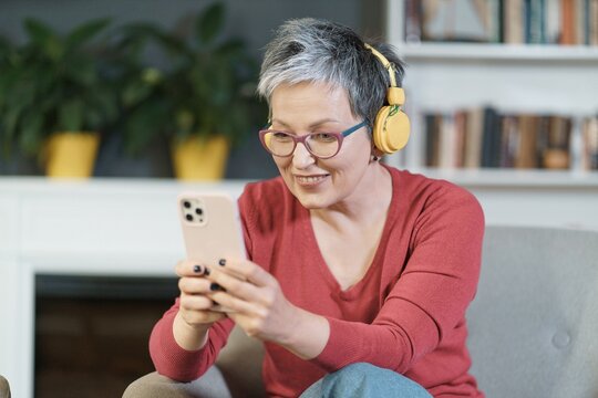 Mature woman wearing pink glasses and yellow headphones smiles while texting on her phone at home. The image depicts technology, communication, and leisure, while the woman's happy expression reflects