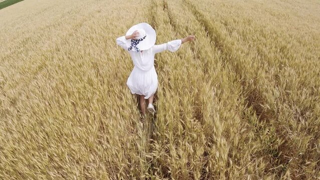 Happy woman in white dress and hat running on wheat agriculture harvest field, slow motion