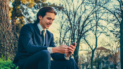 Obraz premium Happy man in a business suit sets outdoors and using a smartphone. Portrait of the young man in a business suit with mobile phone, in the park. Smiling businessman in city looking to a cell phone.