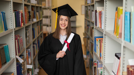 Happy young woman in graduate gown holding diploma in the library. 