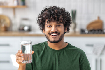 Young Asian man holding a glass of clean fresh water looking at the camera and smiling standing at...