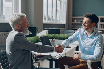 Two confident businessmen shaking hands and smiling while sitting at the office desk together