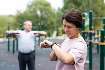 Mature woman in sportswear looks at fitness watch or at pedometer while exercising in city park