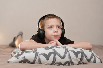 The boy lies on the bed on the pillow  in headphones and watches TV, holding a control panel in his...