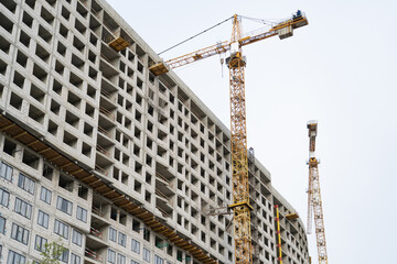 Yellow construction tower cranes and unfinished residential building against grey sky. Housing...