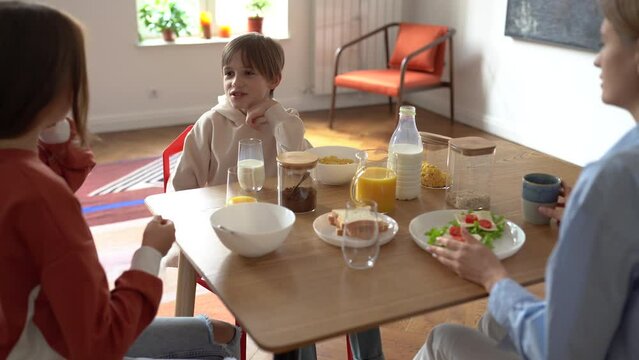 Happy family mother and two kids talking during breakfast, eating together in morning, children sitting at kitchen table with mom enjoying mealtime conversation, spending weekend with mommy at home