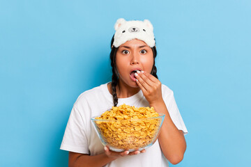 Portrait of enthusiastic asian young woman with pigtails and sleeping mask watching a tv eating corn flakes from the bowl isolated over blue studio background. People lifestyle morning routine concept