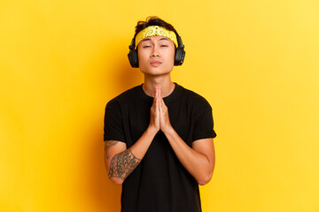 Portrait of satisfied and thankfull ethnic young man with tattoo listens music via wireless headphones enjoys his favorite songs makes gesture with palms folded wears yellow headband and black t-shirt