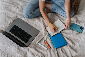 Cropped shoot of girl writing in diary sitting on bed with laptop phone pen, remote learning home. Top view on remote working young woman planning agenda at bedroom. Education, business people. Gadget