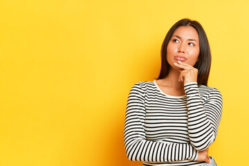 Horizontal shot of pensive Asian pretty woman keeps fingers on a chin looks thoughtfully aside...