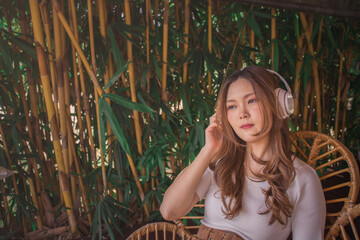 Relaxed asian woman with wireless headphones, listening to meditation music and smiling with pleasure. Joyful young girl wears white t-shirt sitting on chair in cozy botanical garden. lifestyle.