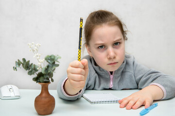 frowning girl sits at a table holding a pencil in her fist and threatening someone with it