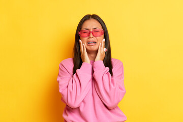 Indoor shot of hesitant brunette woman shrug shoulders put hands on the face cry and doubt while make decision stands puzzled wears modern pink sweater and pink glasses isolated over yellow background - obrazy, fototapety, plakaty