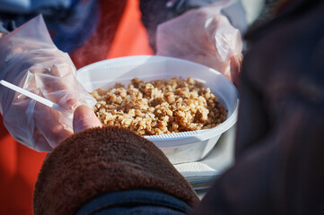 A volunteer passes a plate of food to a starving man on the street. The concept of poverty and hunger. Groats of hot buckwheat porridge. Sunny cold day. Selective focus.