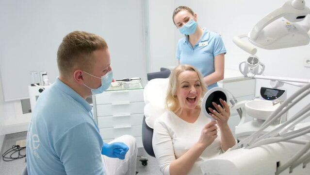 classical dental coloring for determining color of teeth dental clinic doctor hands over mirrors middle aged woman patient she is happy with result smiles shows thumbs up laughs and gives mirror