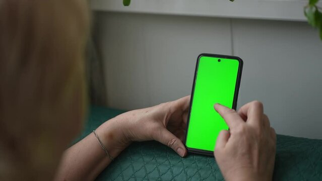 Use green screen for copy space closeup. Chroma key mock-up on smartphone in hand. Woman holds mobile phone iPhone and swipes photos or pictures left indoors of cozy home