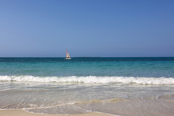 Blue waves breaking on white sand, tropical beach with sailboat in ocean. Background for holidays on a paradise nature