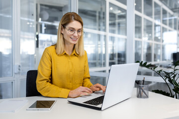 Happy and successful business woman working inside office with laptop, female worker in yellow...