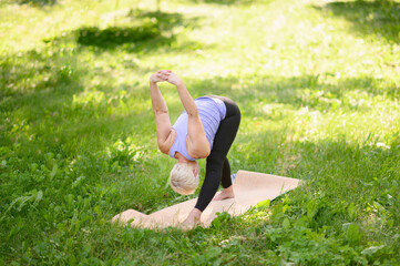 Fototapeta premium Middle aged woman doing yoga in sportswear outdoors in city park in Standing Forward Bend .Uttanasana pose. The concept of stretching, pilates, healthy lifestyle. 