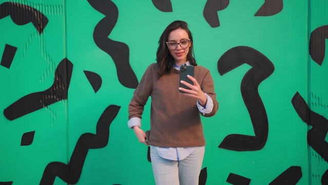 Woman taking a selfie on smartphone camera outside. Young adult female person in glasses with leopard print doing selfie photo on mobile phone camera smiling showing tongue on outdoor