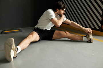 Athletic handsome male football player with muscular strong body training at gym, preparing himself for game, stretching legs and shoulders, sitting on floor, breathing slowly and deeply