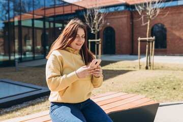 Beautiful woman in casual clothing using her smart phone while sitting on the bench on city street. Happy Young Middle Eastern woman sitting on a bench after college classes. Urban lifestyle concept