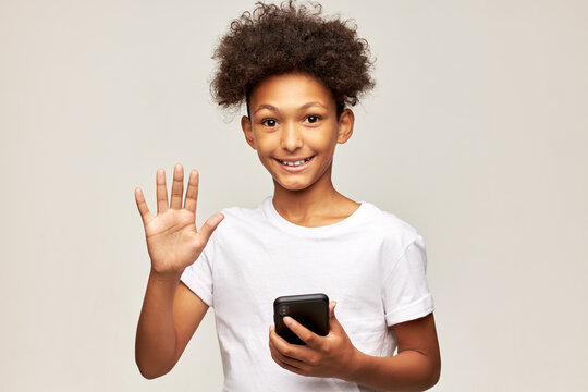 Studio portrait of happy friendly kid boy of 12 waving hand at camera talking to his classmates on smartphone using video chat and online phone application, posing against gray background