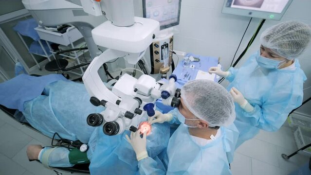 Ophthalmological surgery specialist sits in front of microscope performing operation. Low angle view on the female doctor conducting high-precision surgery. Femto Laser Assisted Cataract Surgery.