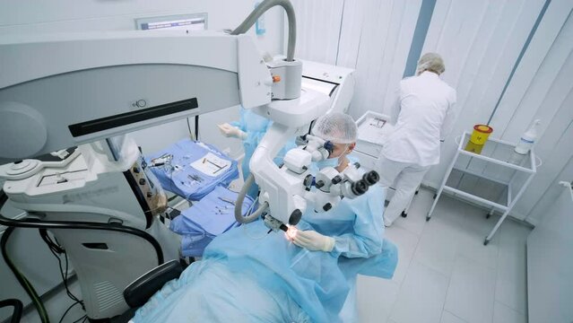 Ophthalmological surgery specialist sits in front of microscope performing operation. Low angle view on the female doctor conducting high-precision surgery. Femto Laser Assisted Cataract Surgery.