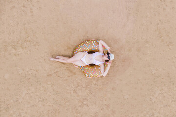 Fototapeta na wymiar Aerial view of a young woman in a swimsuit, hat, sunglasses on an inflatable big donut on the sand on the beach. The concept of rest, relaxation, resorts, water activities, the danger of sunburn.
