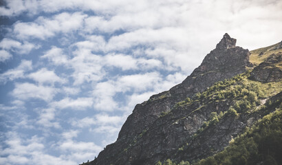 Naklejka premium Mountain landscape with a rocky slope overgrown with vegetation against a cloudy sky in the mountains, summer landscape in the mountains
