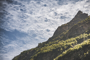 Naklejka premium Mountain landscape with a rocky slope overgrown with vegetation against a cloudy sky in the mountains, summer landscape in the mountains