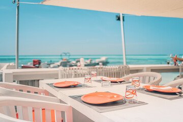 Relaxing outdoor restaurant at the beach. Table setting at tropical beach restaurant. Elegant white wooden tables, chairs under beautiful soft sunset sky, sea view. Luxury hotel or resort restaurant
